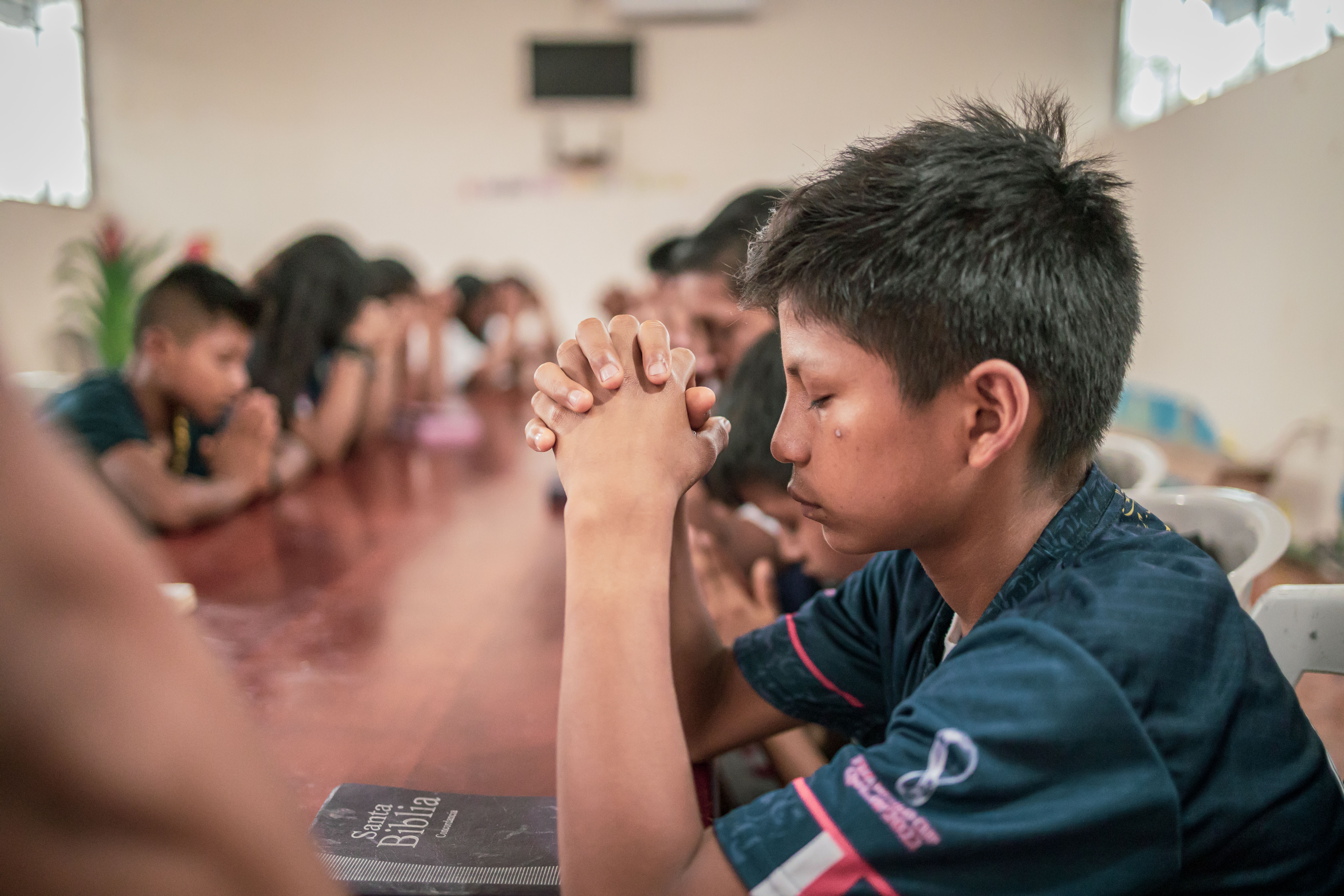  Young boy sits at a table with his friends with his hands clasped in front of him and his eyes closed.