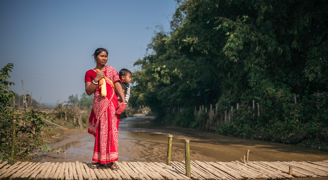 A mom wearing a bright red dress holds her baby in a sling while standing on a wooden bridge over water.