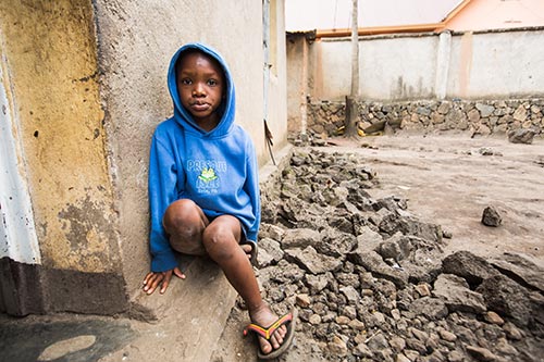 A boy wearing a blue hooded sweatshirt sits against a wall