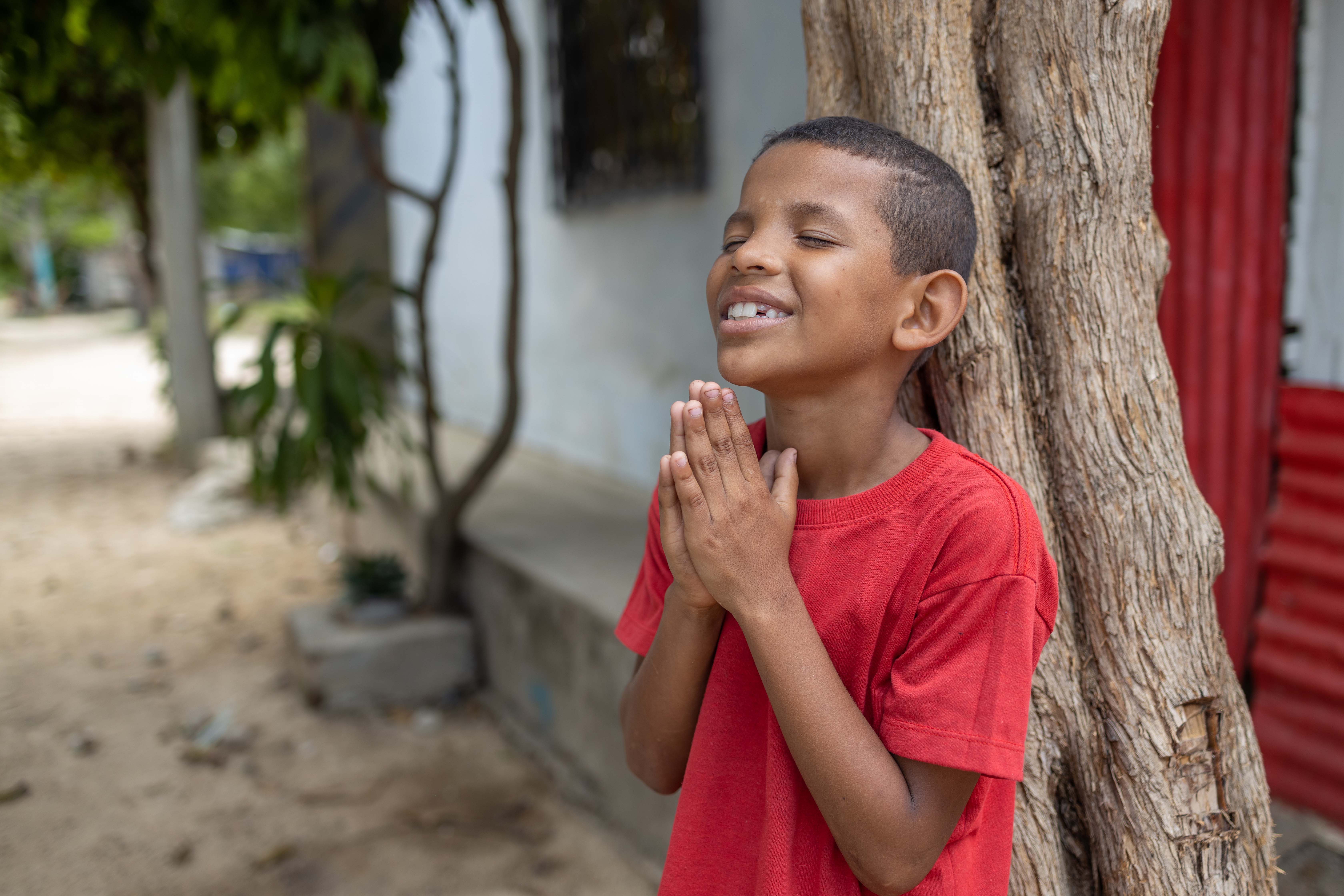 Young boy wearing a red shirt leans on a tree with his eyes closed and hands clasped together as he smiles and prays.