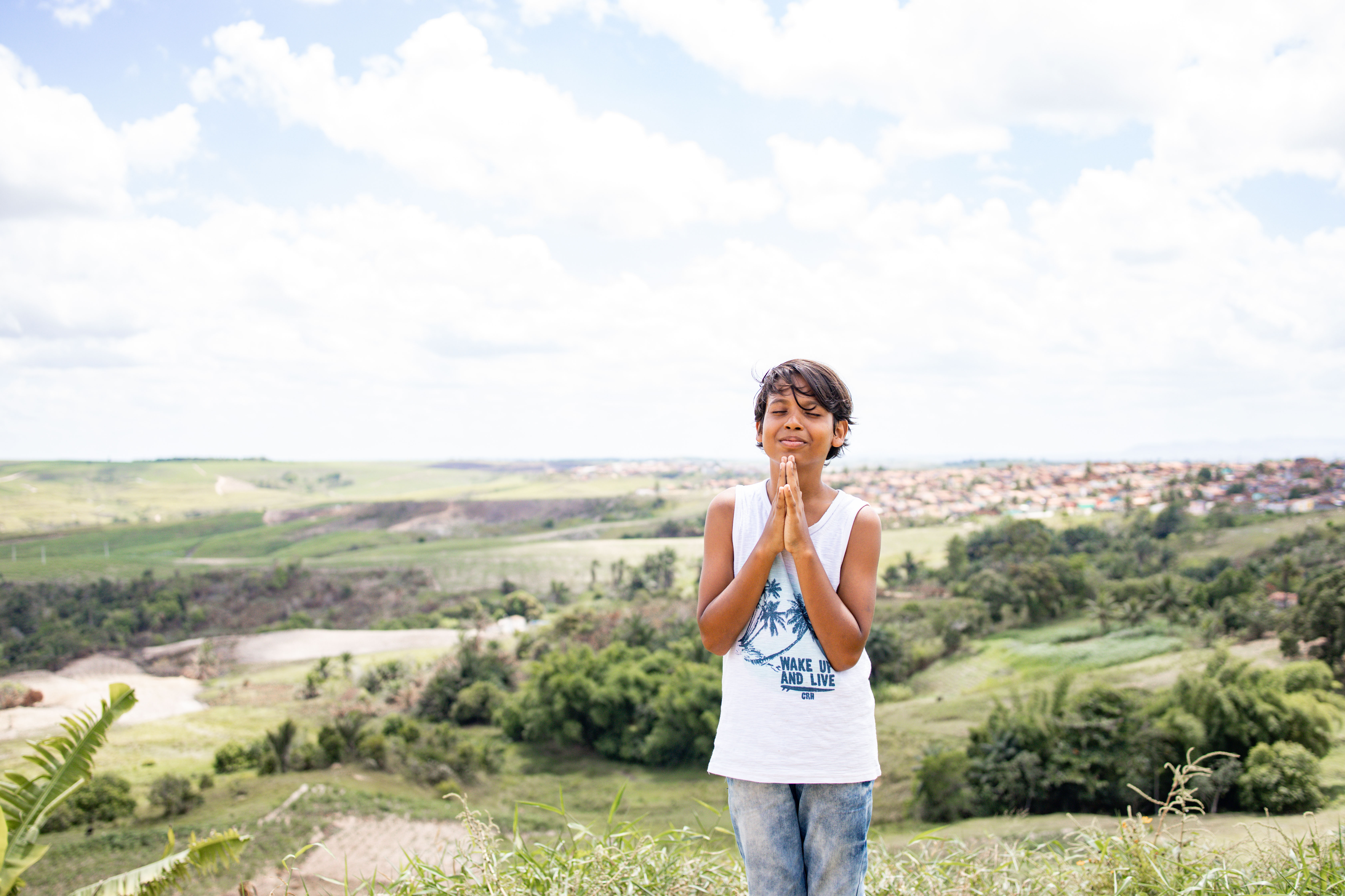 Young boy with dark hair and wearing a white tank top stands with his eyes closed and hands together in front of his face. The background is a beautiful green landscape with a bright blue sky filled with clouds.