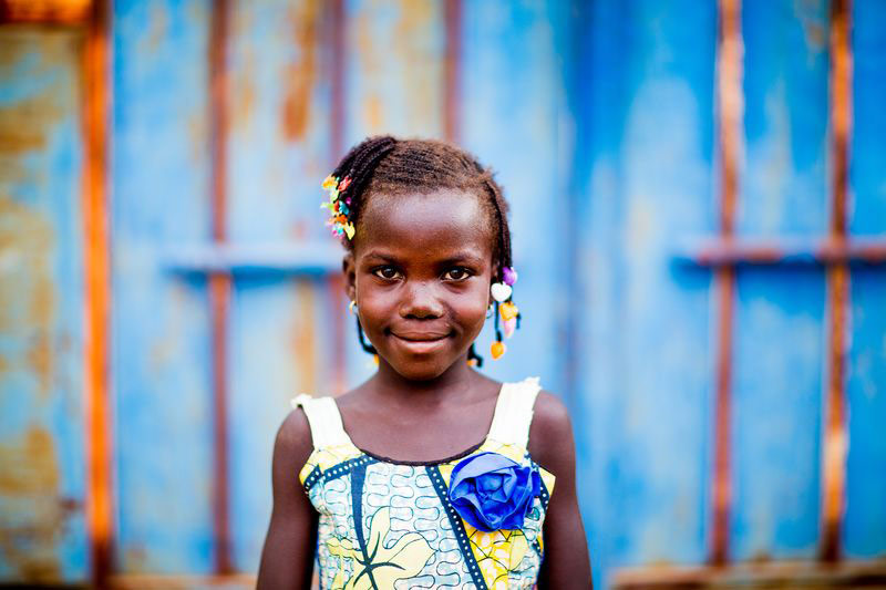 A young girl stands smiling in front of a bright wall