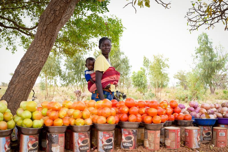 A mother carries her young child on her back as she sells vegetables to generate income for her family