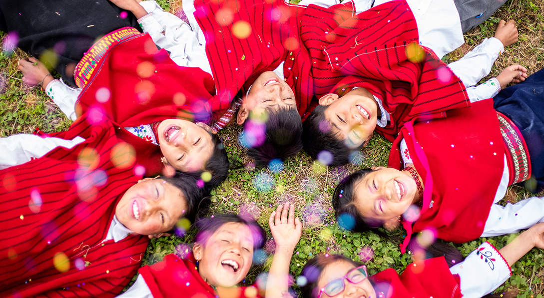 A group of children lay on the ground and smile for the camera while confetti falls.