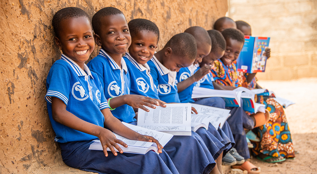 Image shows a group of children wearing blue school uniforms sitting on a concrete step and reading their schoolbooks.