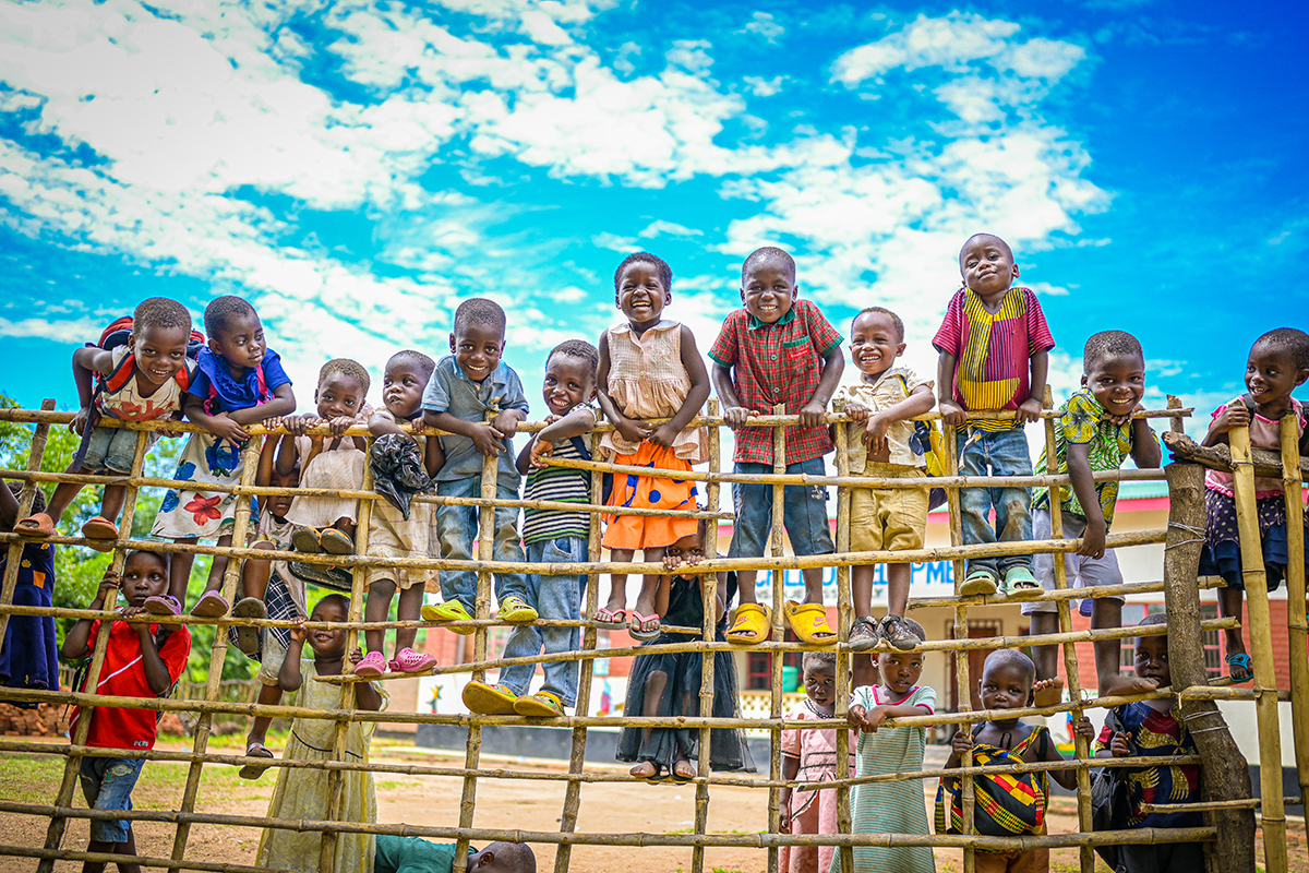 A group of children stand on a fence in front of a bright blue sky and smile for the camera.