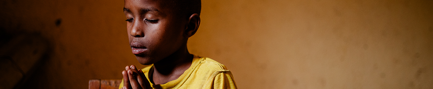 Young boy wearing a bright yellow shirt sits in a chair with his eyes closed and hands pressed together as he prays.