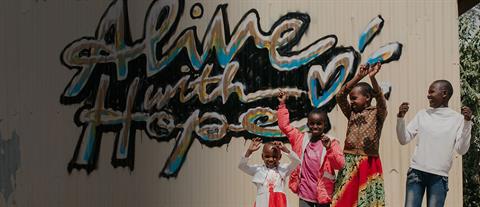 Four children raising their hands in the air in front of a shed with alive with hope painted on it