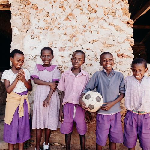 Five children wearing pink and purple uniforms stand in front of a clay home and smile for the camera.