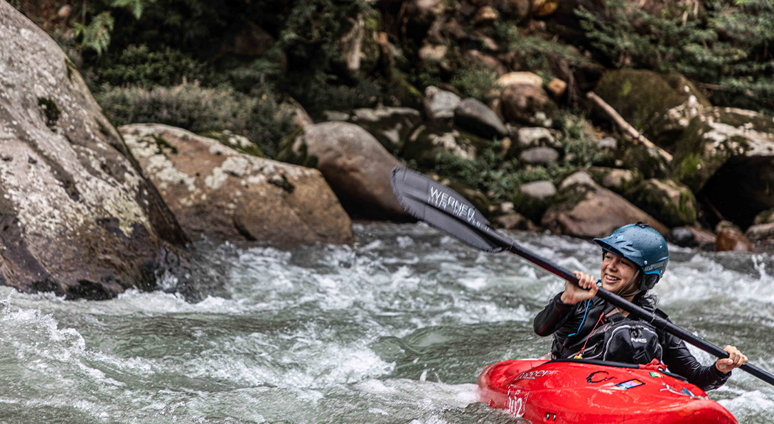 Rafaella sits in her bright red kayak and lifts her paddle up into the air while floating down rapids in the Amazon, Ecuador.