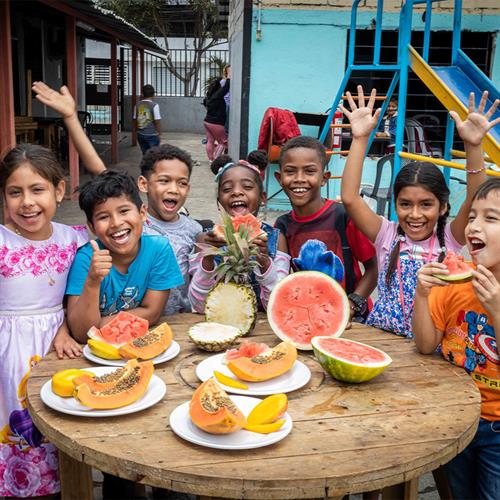 Seven smiling children stand outside around a circular wooden table full of sliced watermelon and cantaloupe. A blue building and slide are in the background.