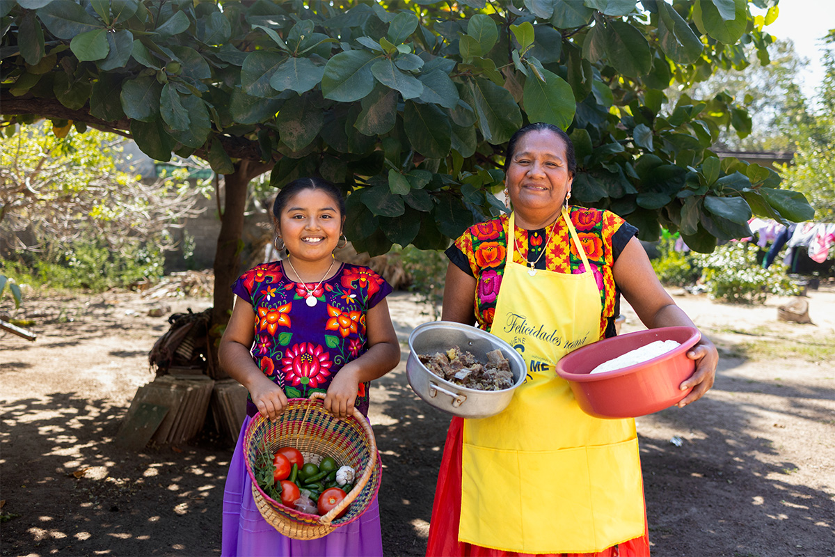 A young girl and her mother stand outside in front of a tree holding pots of fresh veggies and grains. They are wearing brightly colored floral outfits and smiling at the camera.