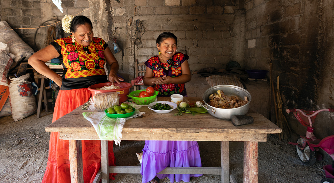 Young girl stands next to her mother behind a wooden table that's covered in pots veggies and other ingredients. Both are wearing floral tops and brightly colored skirts.