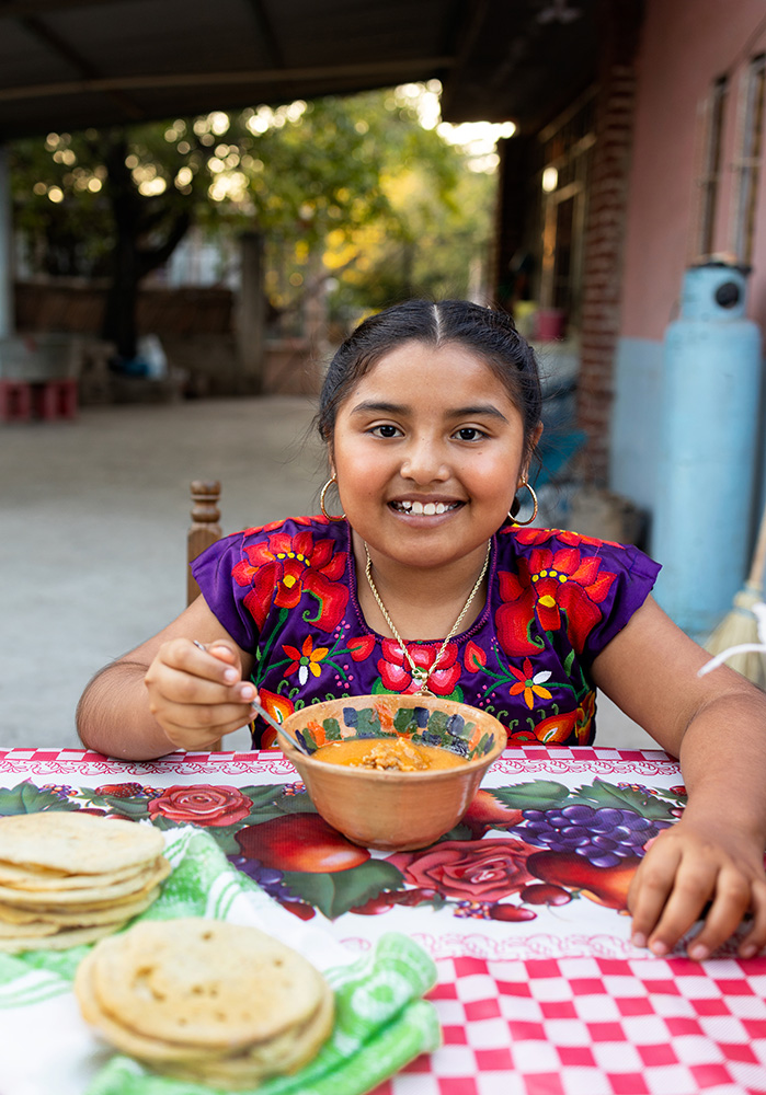 Young girl sits at a fabric-covered table outside with a bowl of bright yellow stew. She wears a colorful floral top and smiles at the camera.