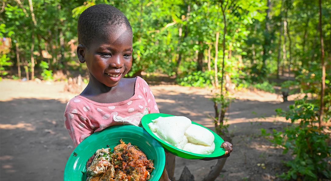 A little girl wearing a pink shirt stands outside, holding two green plates. One plate has an omelet with veggies and the other has round white patties made from corn flour.