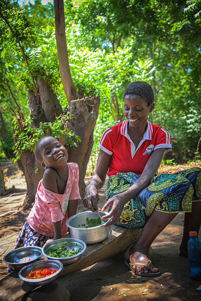 A little girl sits next to her mother on a low wooden bench outside. They are smiling and preparing fresh veggies in metal dishes in front of them.