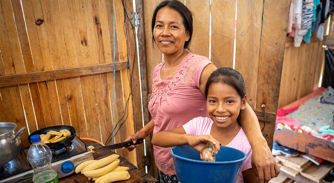 Inside a wooden home, a mother stands with her arm around her young daughter while chopping plantains. The daughter holds a blue bowl full of fresh fish, and both smile at the camera.