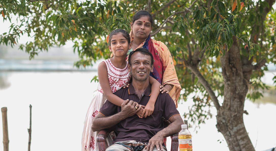 A young girl stands next to her mother in front of a river. She is hugging her father, who sits between them in front of a table of curry ingredients.