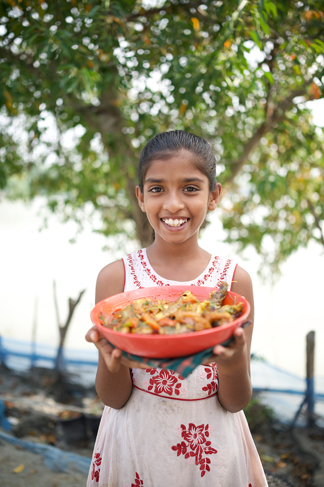 A young girl stands outside in front of a river, holding up a plate of yellow curry and smiling at the camera. She wears a white dress with a red pattern.