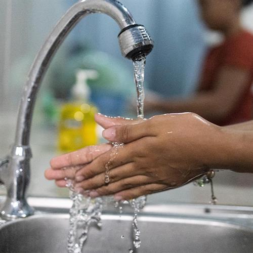 A child’s hands rub together under running water from a sink. More children wash their hands at sinks in the background.