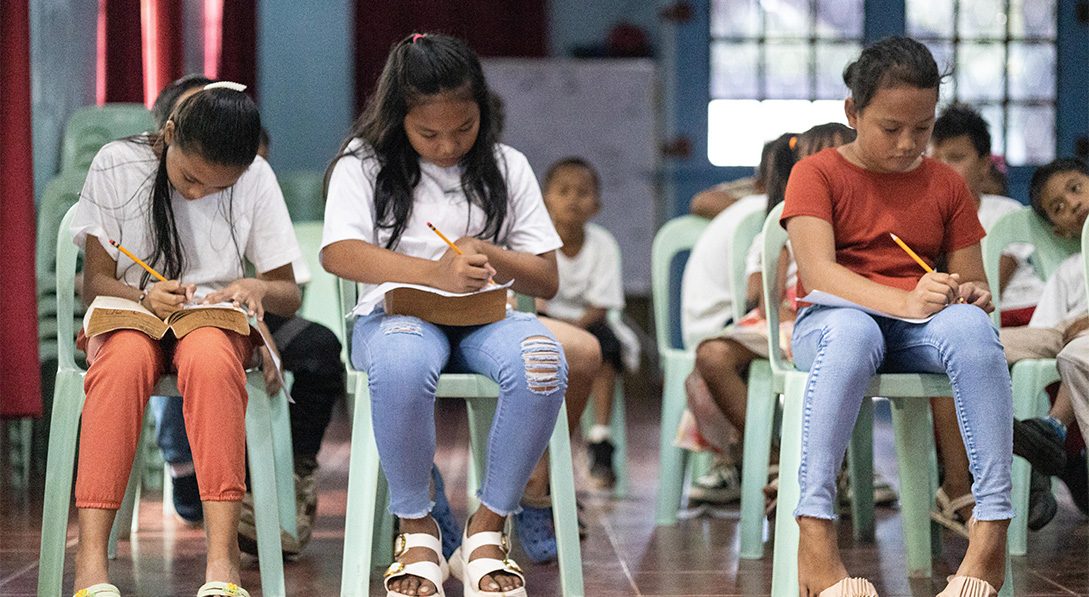 A large group of children sit in green plastic chairs in a classroom and write with pencil and paper on their laps.