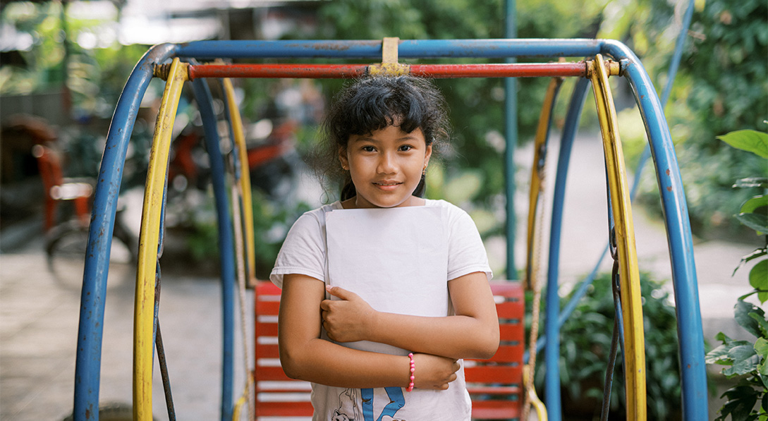 A young girl wearing a white shirt stands in front of a colorful swing outside. She hugs a letter and looks at the camera.