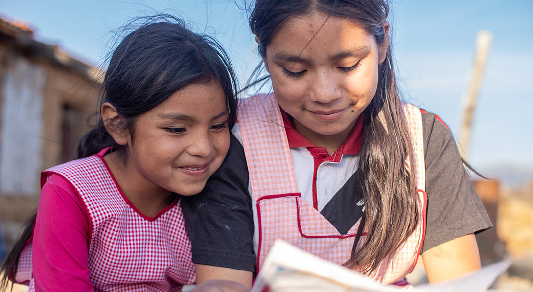 Two little girls wearing pink aprons sit shoulder-to-shoulder. They read a letter with smiles on their faces.