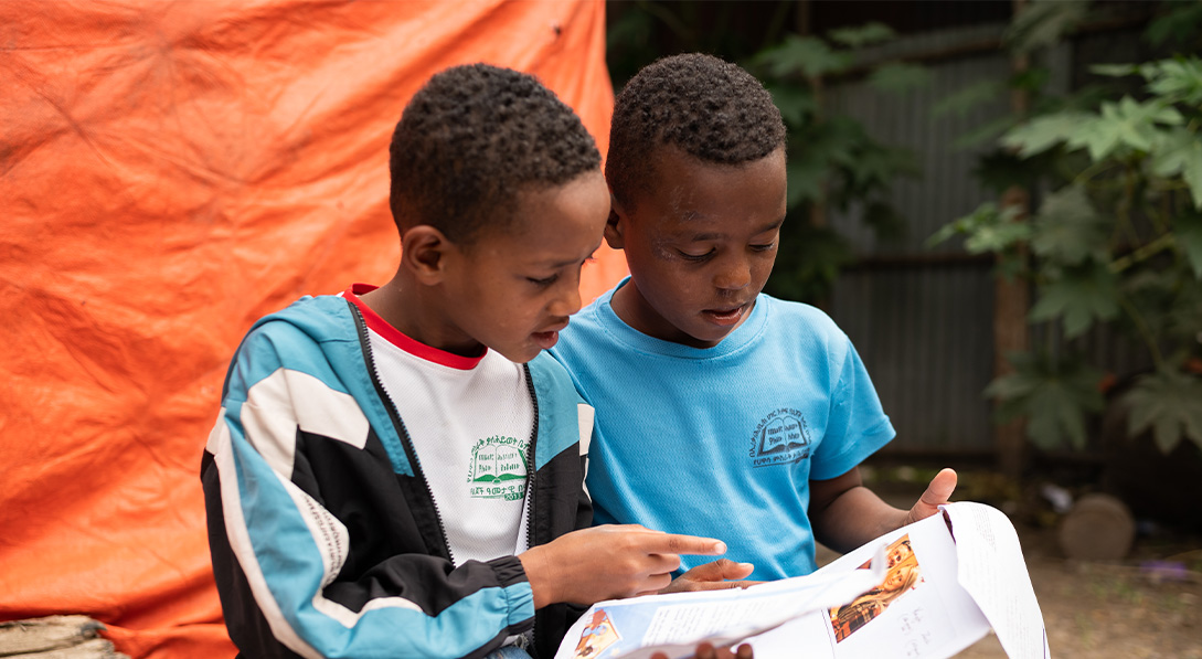 Two little boys read letters together while sitting on a crate outside an orange tent.