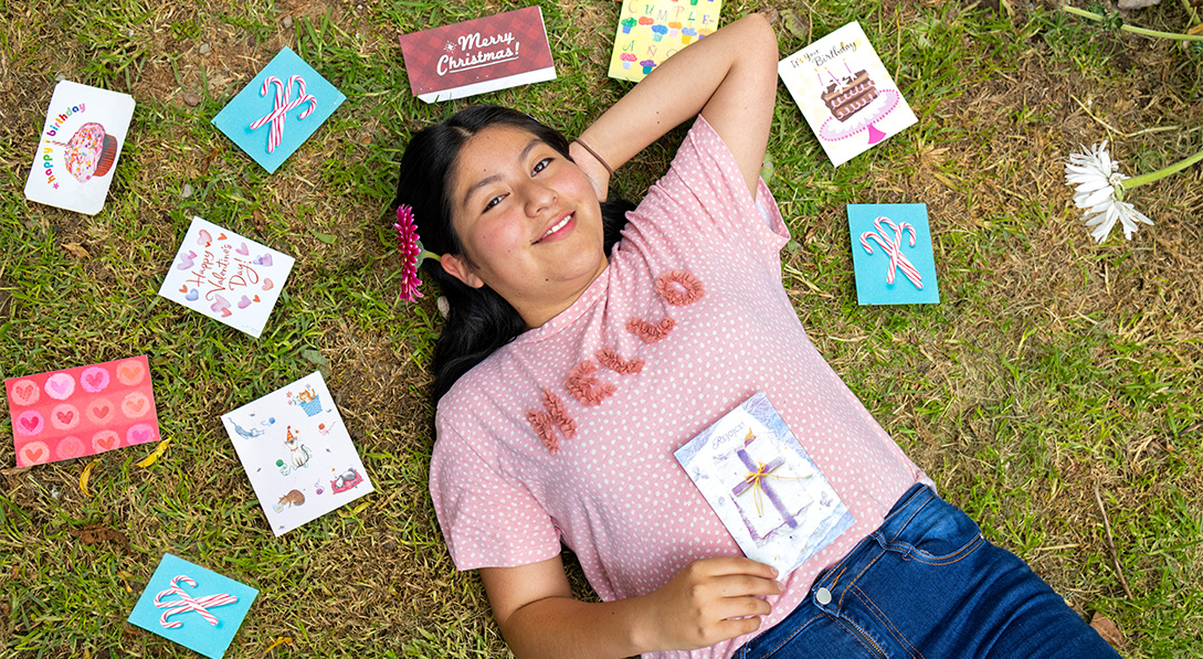 A teenaged girl wearing a pink shirt and jeans lies in the grass, smiling up at the camera. She is surrounded by greeting cards from her sponsor.