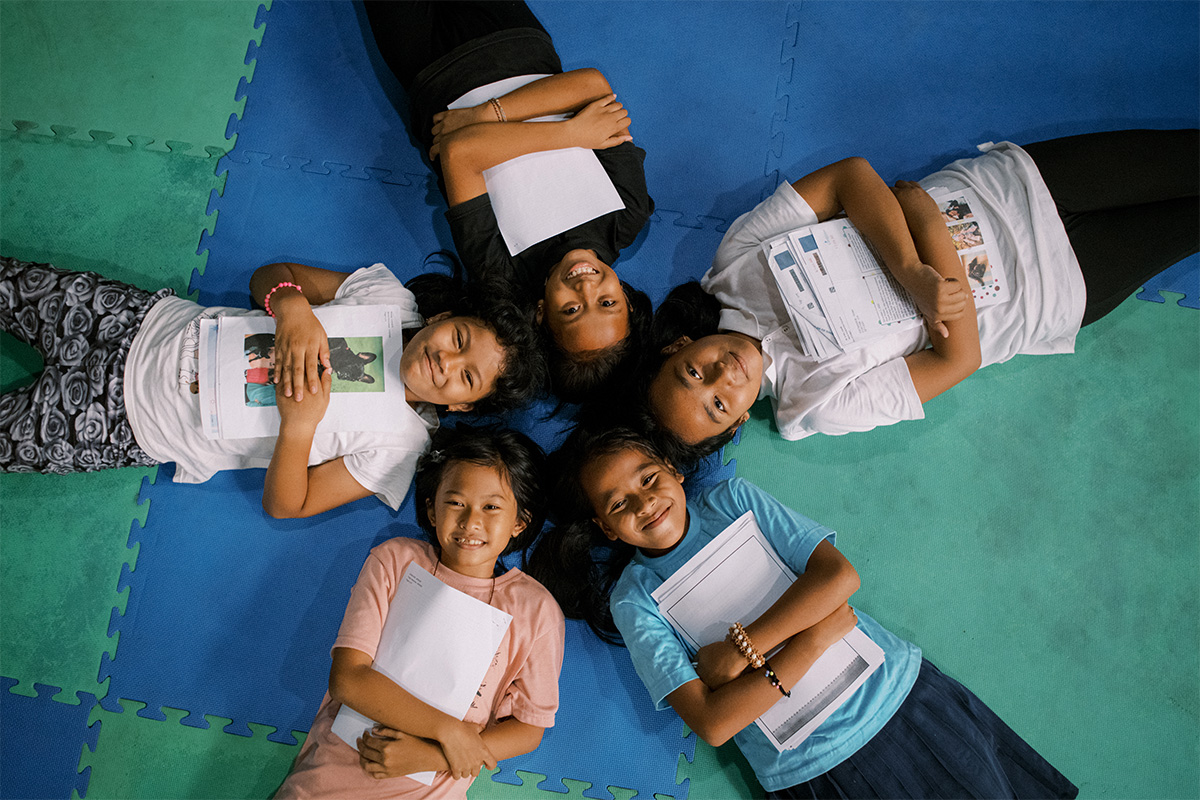 Five young children lie on a colorful mat in a circle. They are hugging letters and smiling up at the camera.