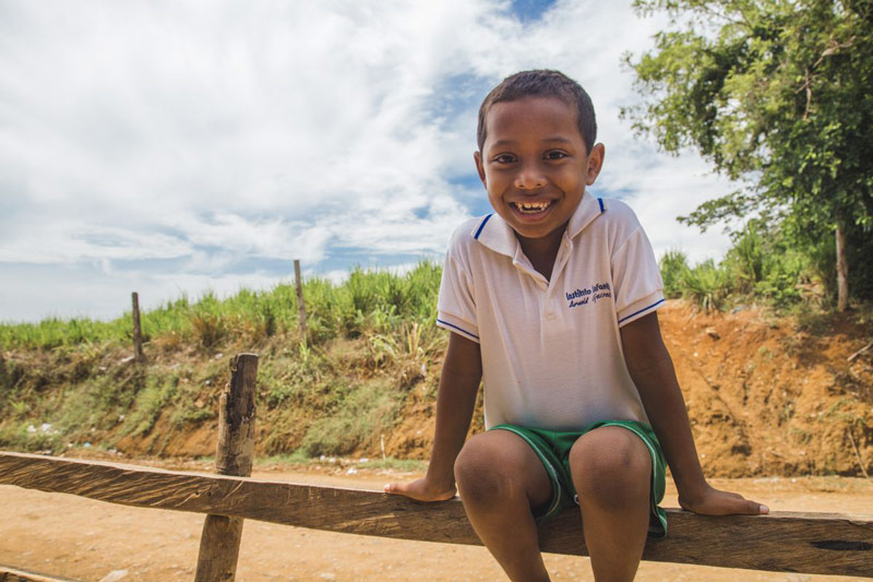 A boy smiles while sitting on a fence near his home