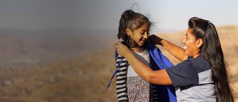 A woman kneels down to place a blue cape on a young girl