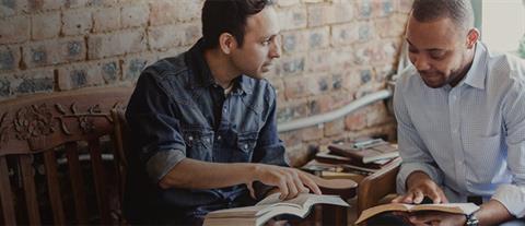 Two men sitting in separate chairs each holding a book