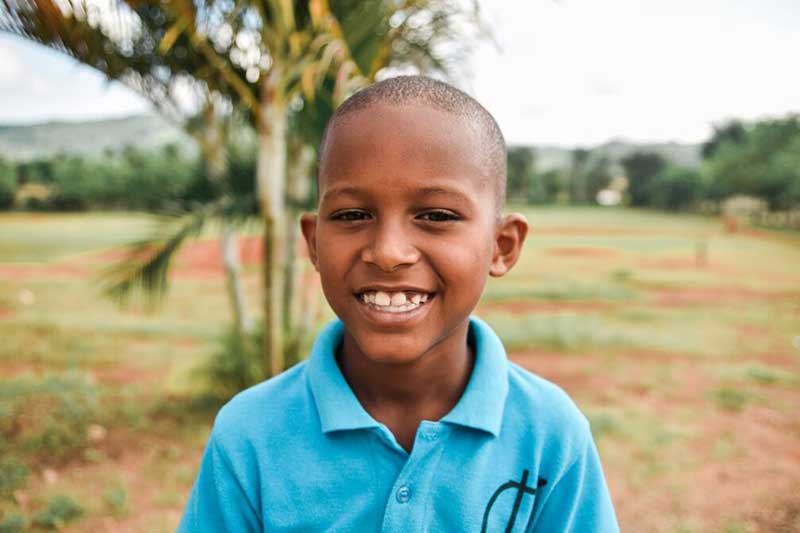 A boy smiles in front of a baseball field