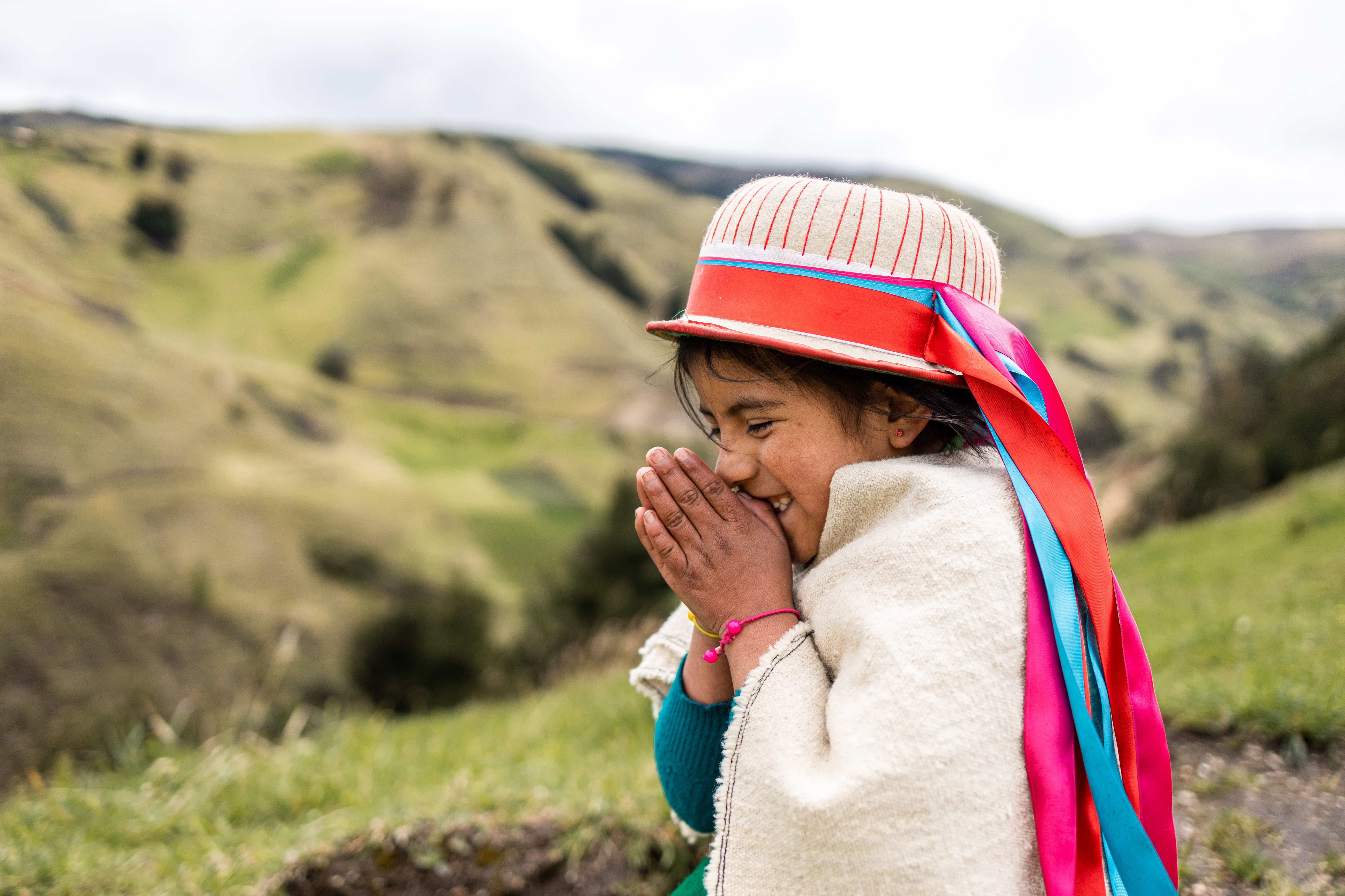 Young girl wearing a hat with brightly colored ribbon holds her hands together in front of her face and grins with her eyes closed.