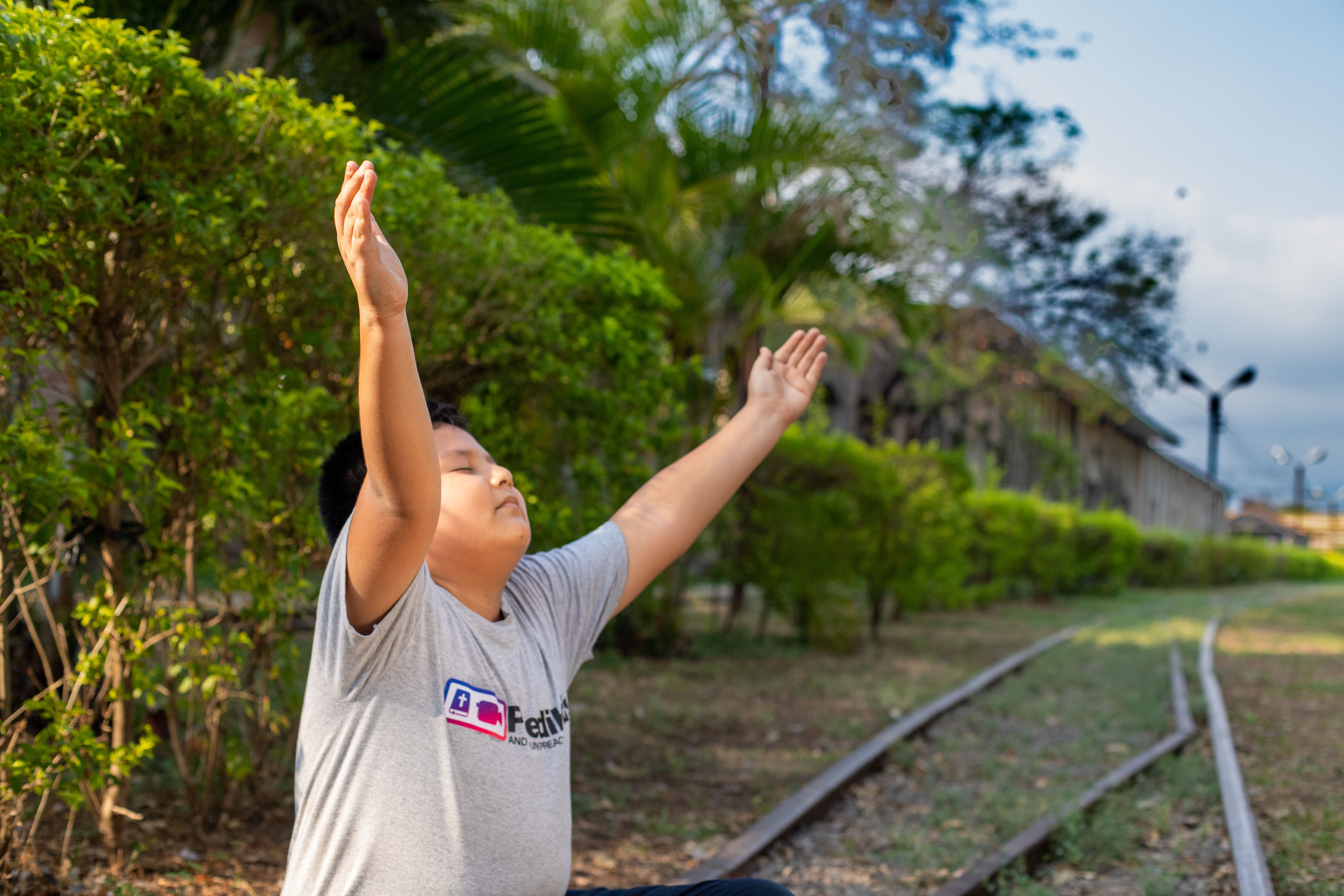  A young boy sits on train tracks with his eyes closed and hands lifted in the air.