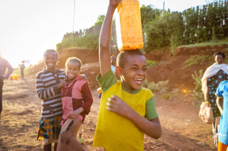 A boy laughing while carrying a water jug on his head