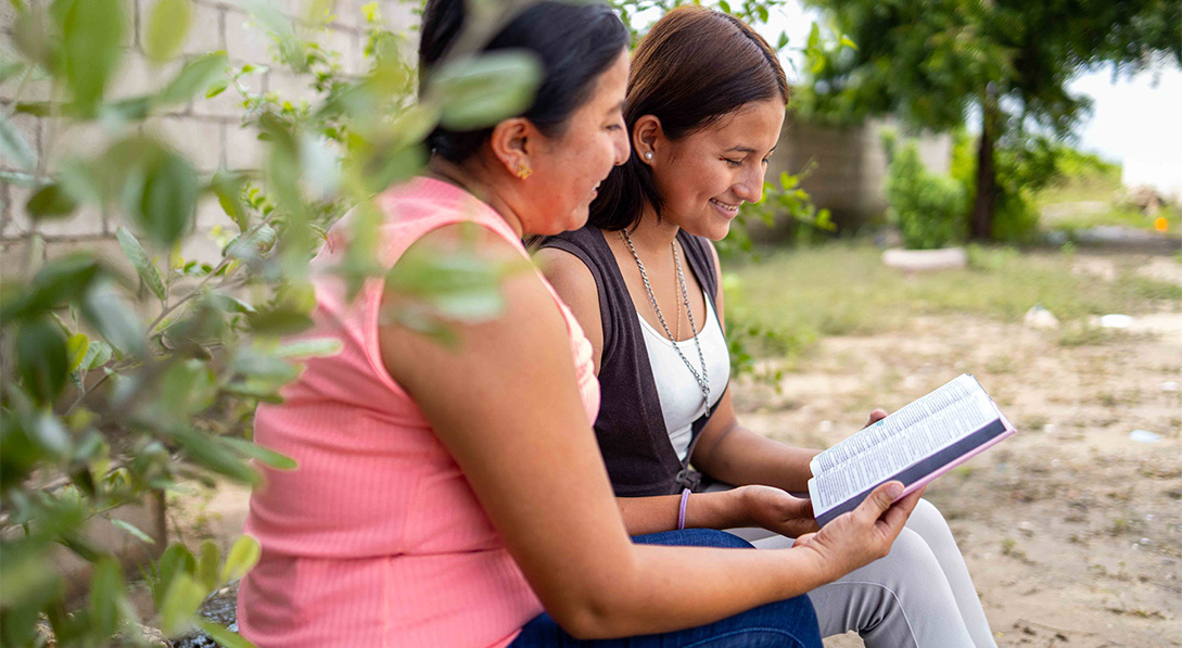 Two women sit next to each other in front of a brick wall while smiling and reading from the Bible.