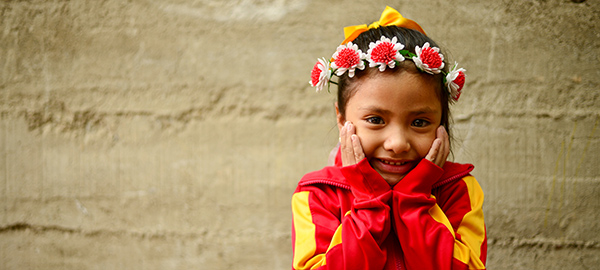 A smiling girl with a flower wreath headband places her hands on her cheeks