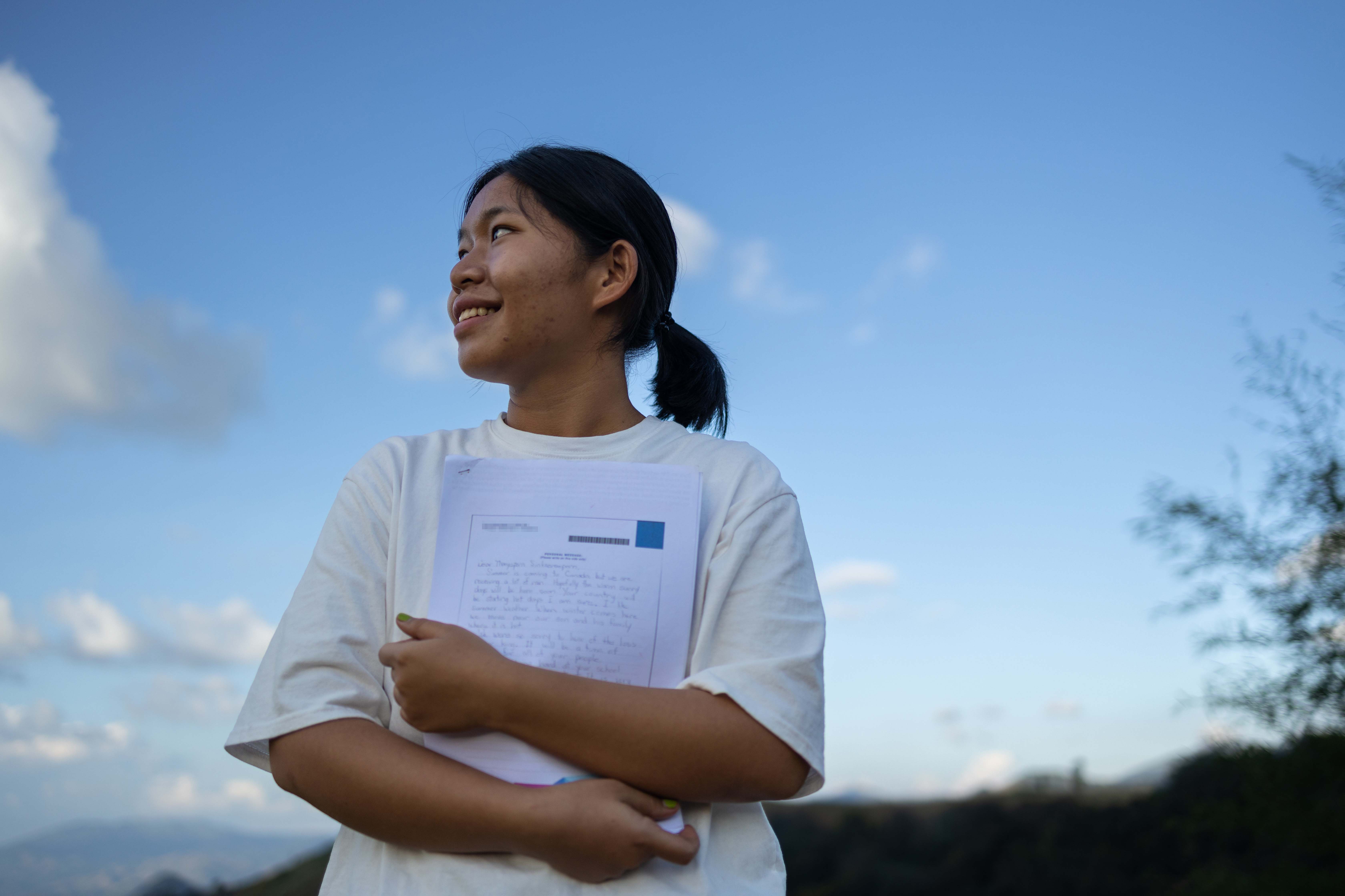 A young woman stands in front of a blue sky and smiles to the right while holding sponsor letters.