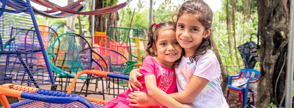 Two girls hugging and smiling at the camera on a colorful patio.