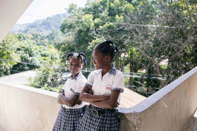 Two girls lean against a wall and smile