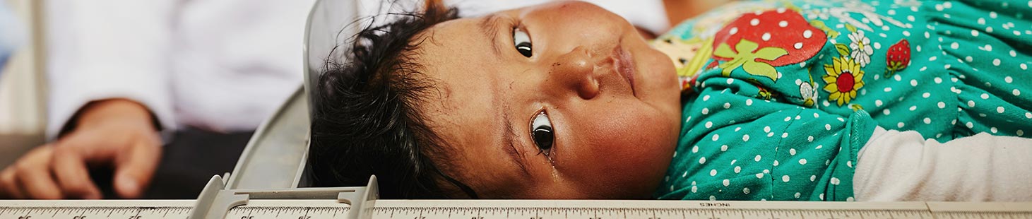 A baby girl wearing green, lies on her back as her height is measured with a stadiometer.