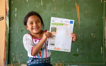 Lizbeth is wearing a white shirt with a floral pattern on it. She is standing in front of a green board in her classroom and she is holding up one of her sponsor's letters.
