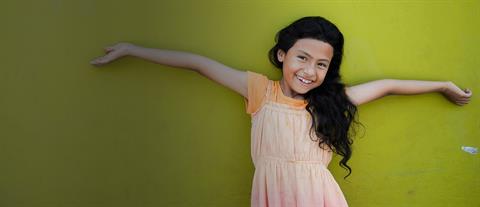 A girl stands in front of a greenish yellow all with her arms spread wide