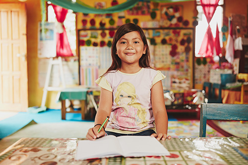 A young Indonesian girl holds a pencil and sits at a table with an open notebook on it