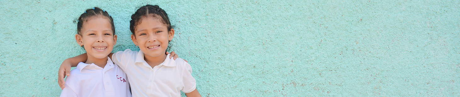 two young girls in white shirts stand in front of a light blue wall with their arms placed around each other's shoulders