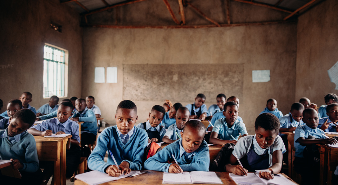 Many children wearing school uniforms sit inside of a classroom, writing in the notebooks on their desks.