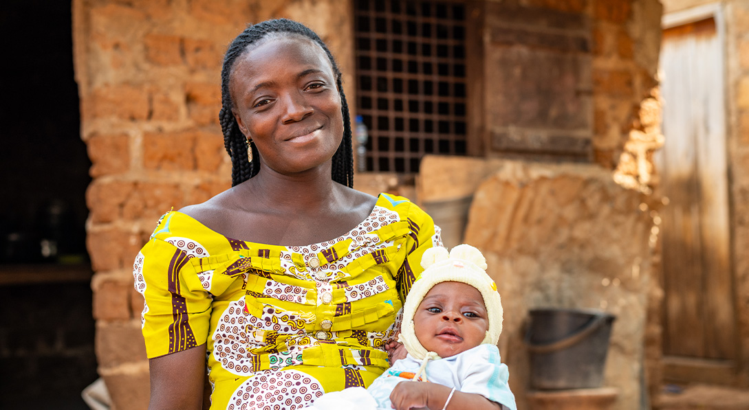 A young woman in a bright yellow pattered dress sits with her baby on her lap. They both smile for the camera.