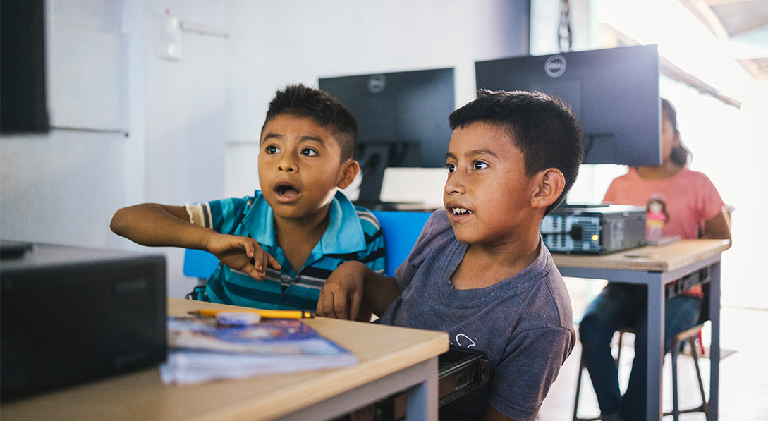 Two young boys sit in front of a computer smiling and looking shocked as they see the screen for the first time.